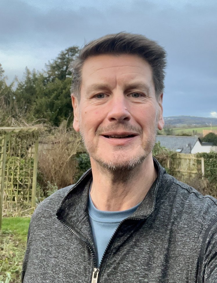 A white man with short greying hair and a beard smiles into the camera. He is outside in the coutryside.