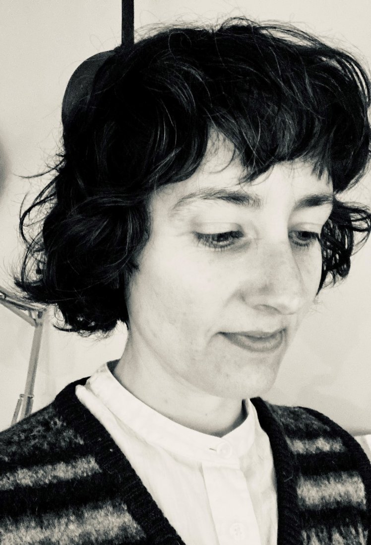 A white woman with short, dark, wavy hair looks down, smiling. She is wearing a knitted top.