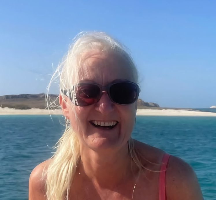 A white woman with long white hair and sunglasses smiling with a sunny seaside view behind her