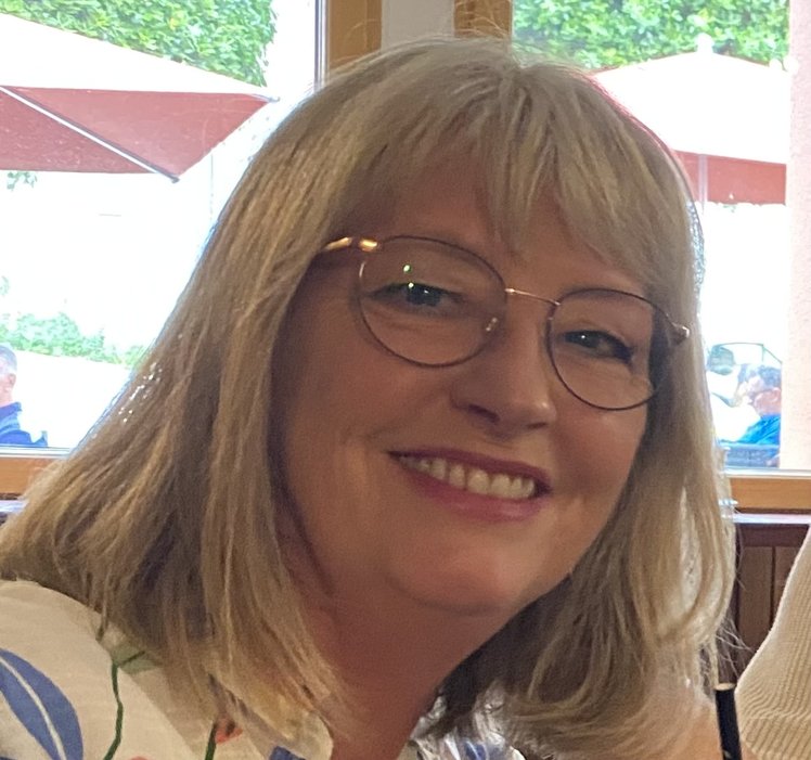 A white woman with blonde shoulder-length hair and glasses smiling broadly with view of people sitting under parasols behind